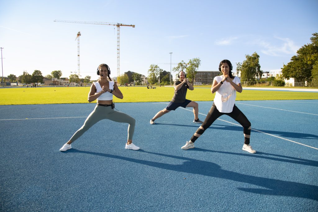 Drei Erwachsene stehen im seitlichen Ausfallschritt auf einem blauen Sportplatz und nehmen ihre Hände an der Brust zusammen. Sie haben alle ein Headset auf.