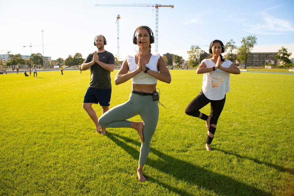 1 Mann und 2 Frauen in Sportkleidung mit Kopfhörern trainieren Yoga auf dem FRANKLIN Sportplatz.