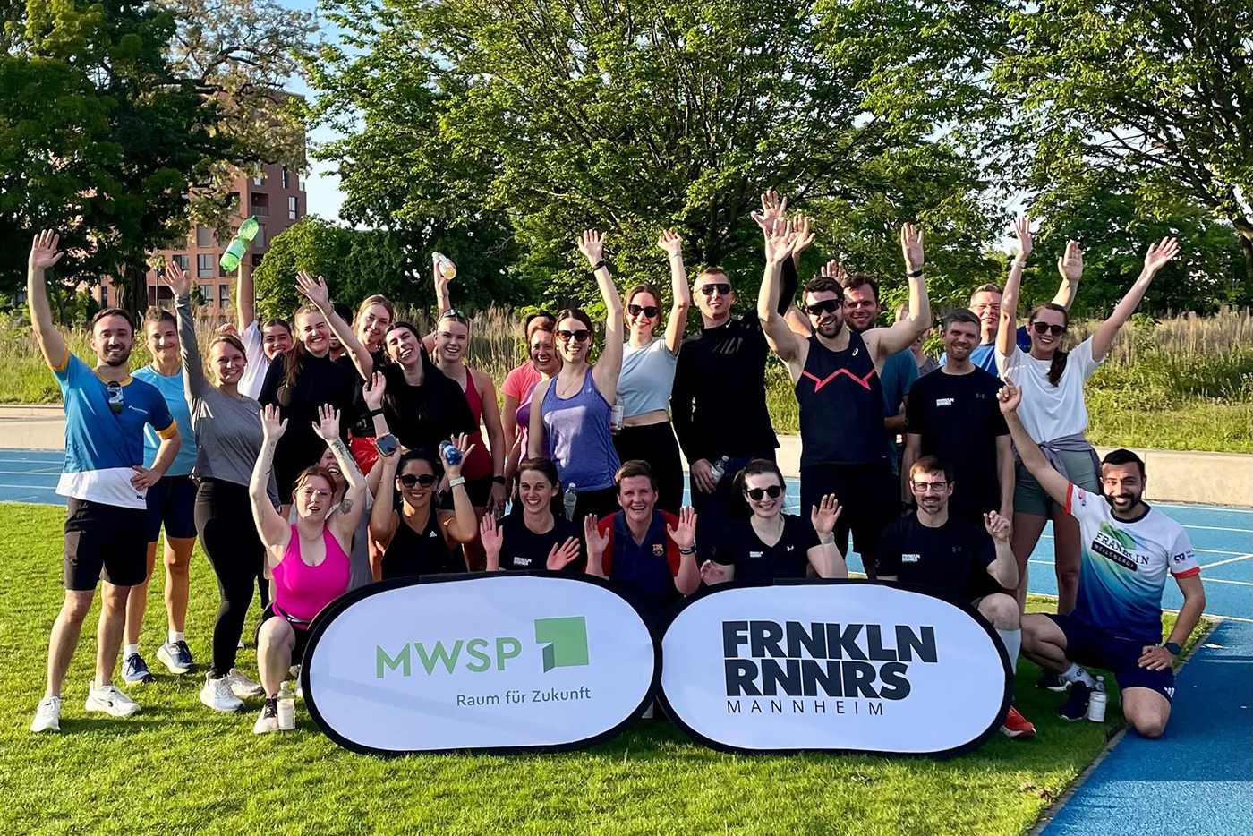 Eine Gruppe von 29 lachenden Läuferinnen und Läufern in Sportkleidung winkt auf einem Sportplatz in die Kamera. Vor Ihnen stehen zwei Banner mit der Aufschrift MWSP, Raum für Zukunft und Franklinrunners Mannheim.