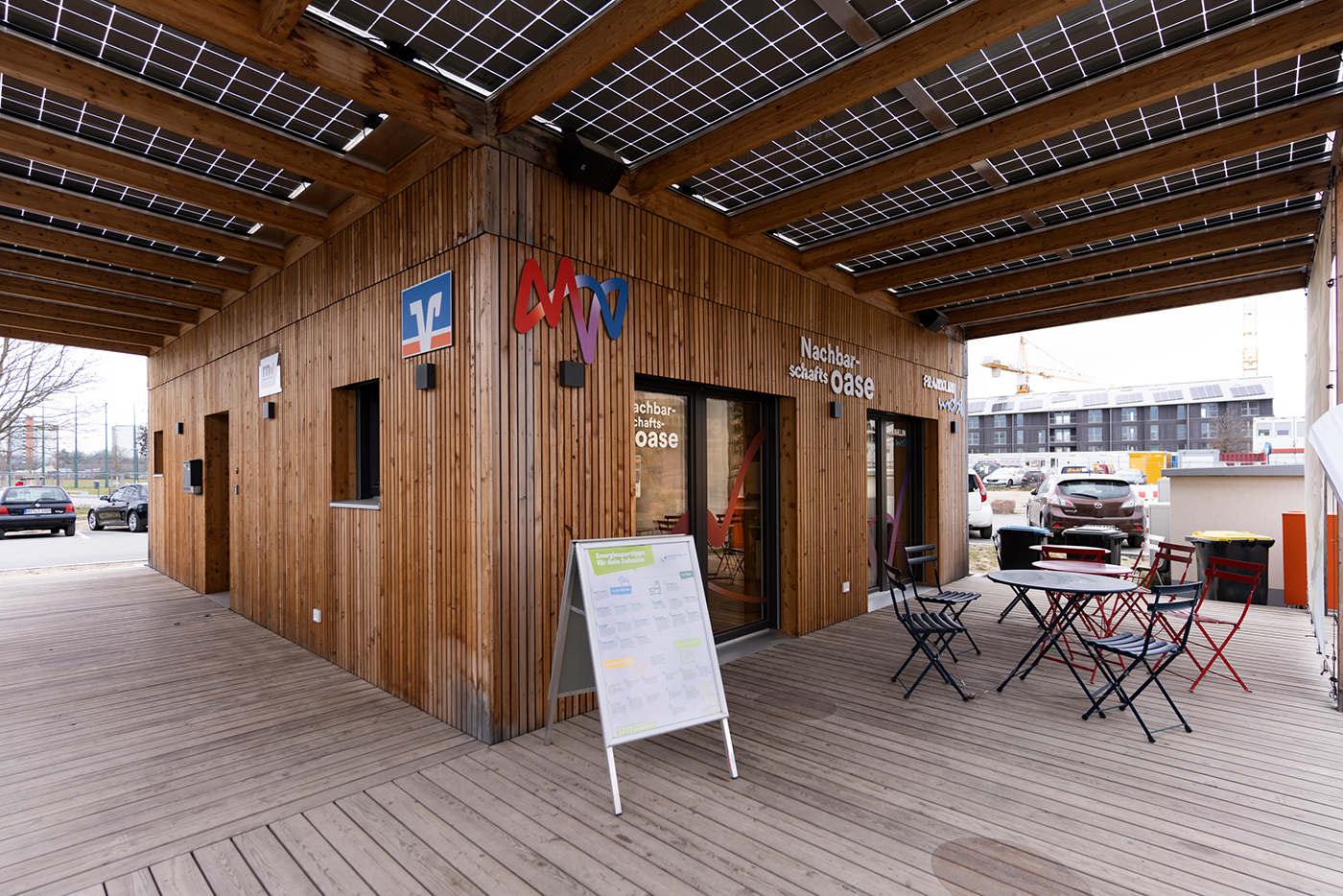 Outdoor-Eingangsbereich mit Terrasse, Dreieckaufsteller, Tischen und Stühlen des quadratischen Holzpavillons der MVV Nachbarschaftsoase. Auf der Fassade sind die Logos von rnv, Volksbank Rhein-Neckar, MVV und FRANKLIN mobil angebracht. 