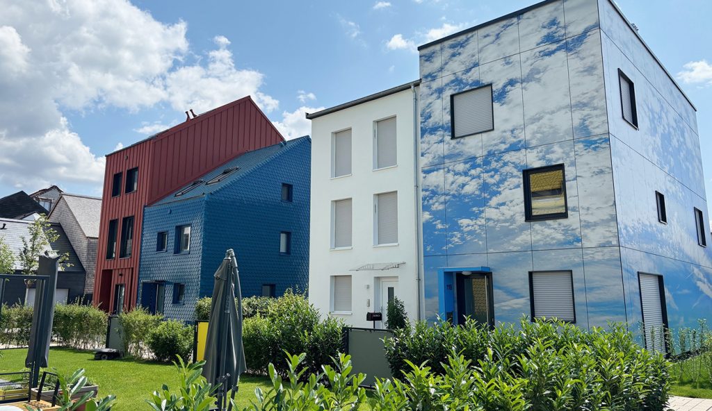 4 bunte Einfamilienhäuser in verschiedenen Formen und Größen. Im Vordergrund ist ein Garten mit Rasenfläche, zusammengeklappten Sonnenschirmen und Abgrenzungshecke sichtbar.