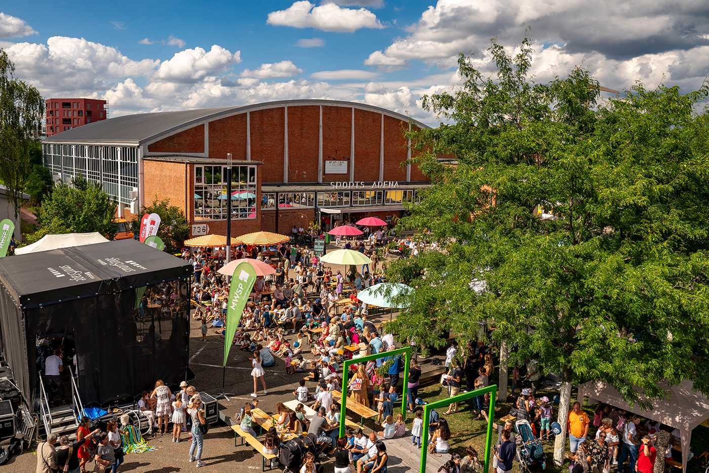 Drohnenaufnahme einer großen Menge Publikum vor einer Bühne. Im Hintergrund sind bunte Sonnenschirme und eine historische Sporthalle mit abgerundetem Dach sichtbar.