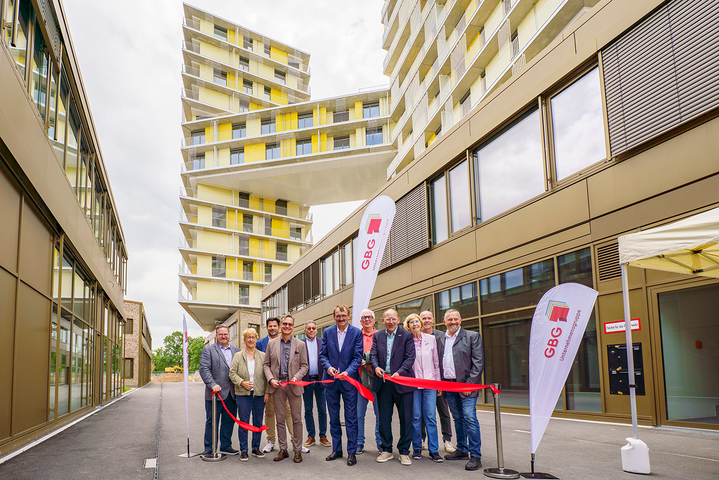 Vor einem Hochhaus in Buchstabenform H schneidet eine Gruppe Menschen mit einer Schere ein Flatterband durch.