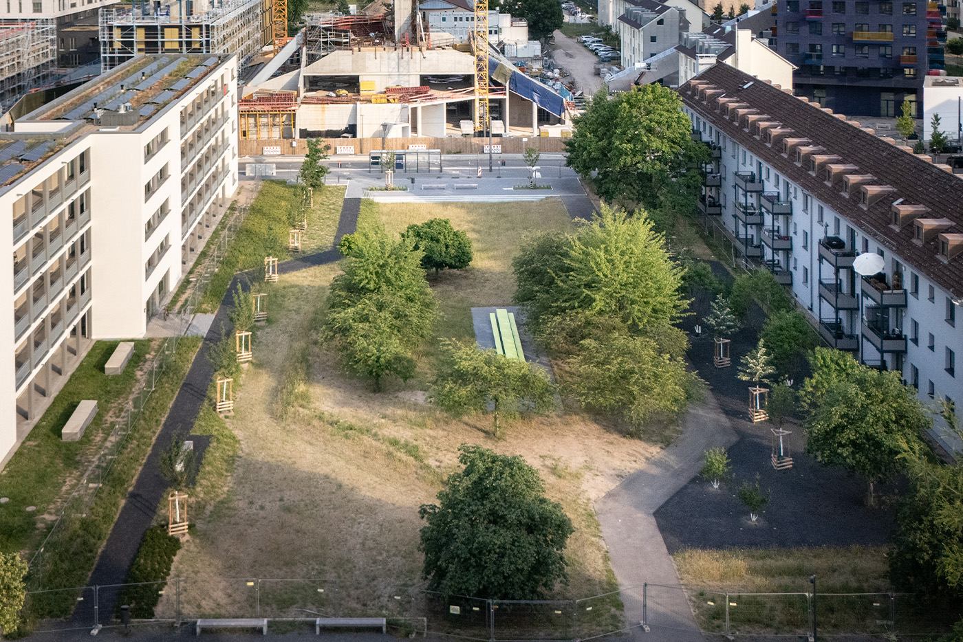 Drohnenaufnahme einer Grünfläche mit Bäumen, Wiese und einer großen Sitzbank in der Mitte. Umrandet wird die Grünfläche mit Wohnbebauung.