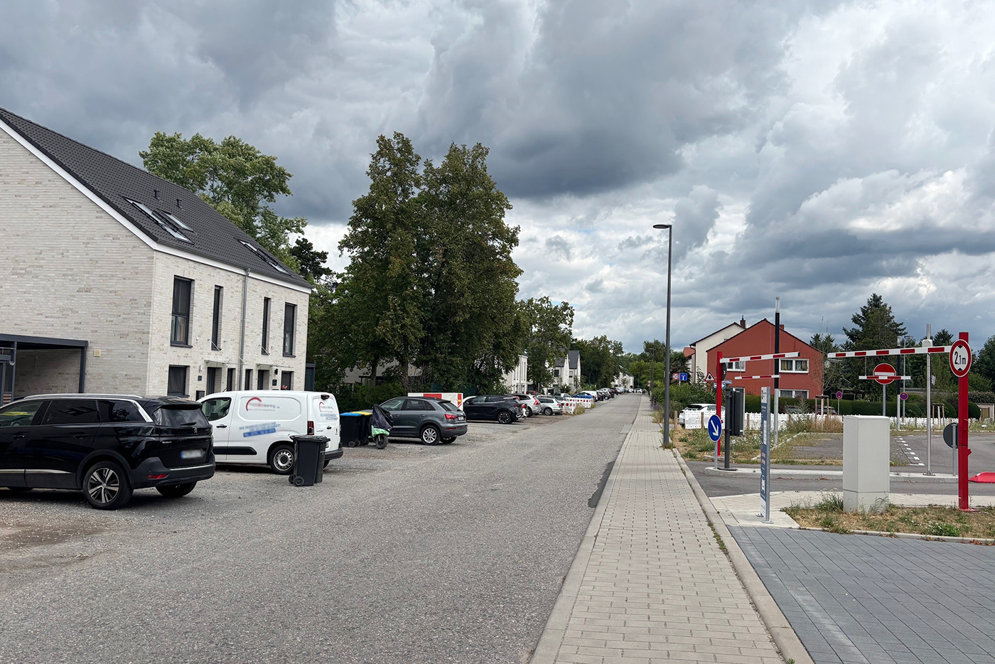 Straßenflucht mit parkenden Autos auf der linken Seite. Im Vordergrund befindet sich auf der rechten Seite eine Einfahrt zu einem öffentlichen Parkplatz.