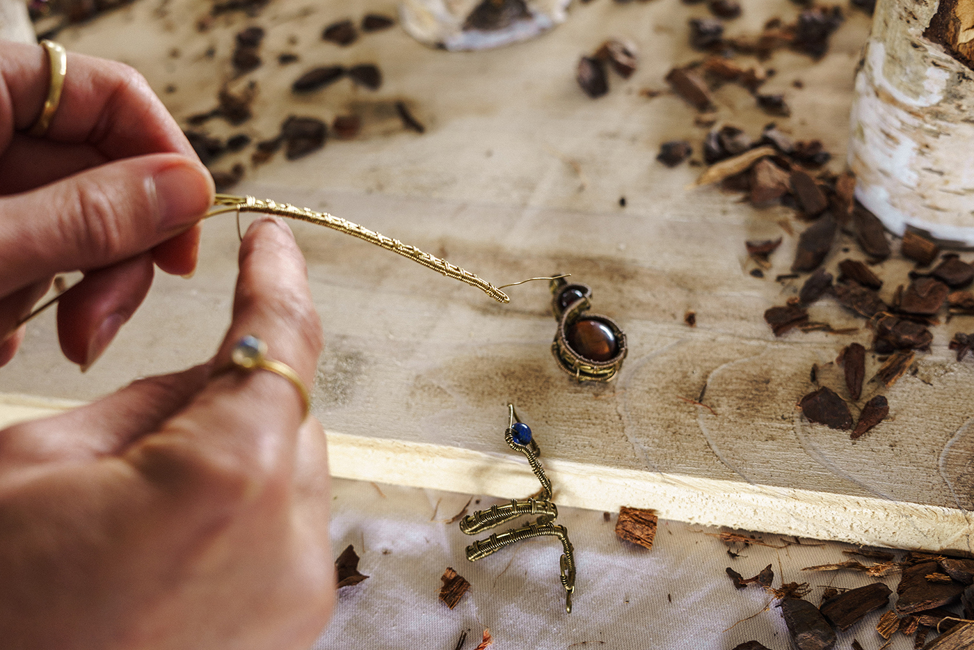 Nahaufnahme von zwei Händen auf einem herbstlich geschmückten Holztisch beim Flechten einer Goldkette, an der als Anhänger ein elipsenförmiger, dunkler Edelstein angebracht ist.