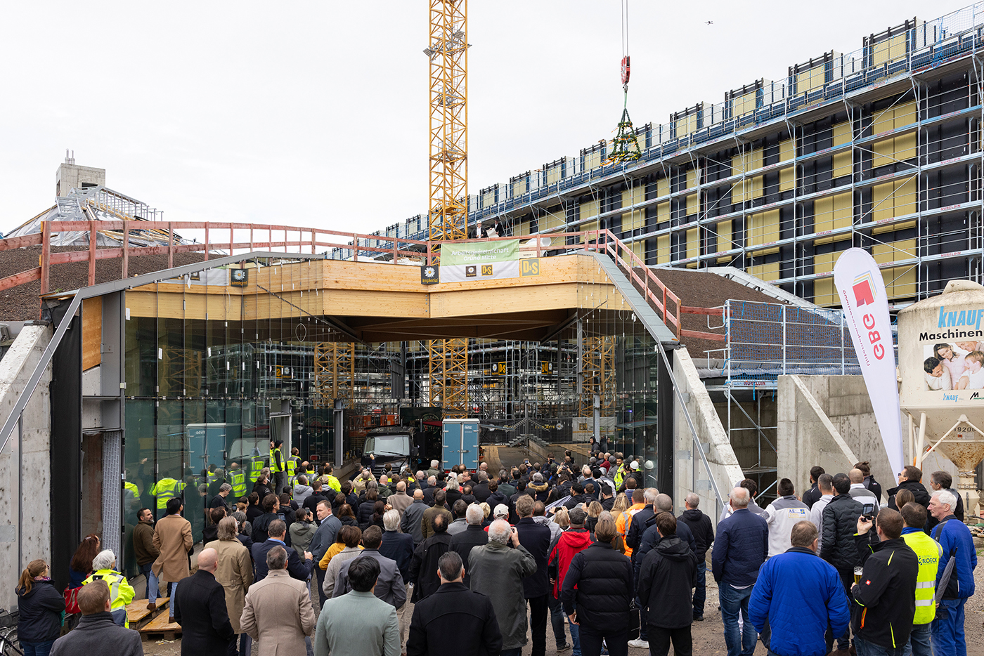 KI-basierte Bildbeschreibung | Eine Menschenmenge versammelt sich auf einer Baustelle, auf der ein teilweise gebauter Glasbau, ein Kran, ein Gerüst und Bauarbeiter zu sehen sind.