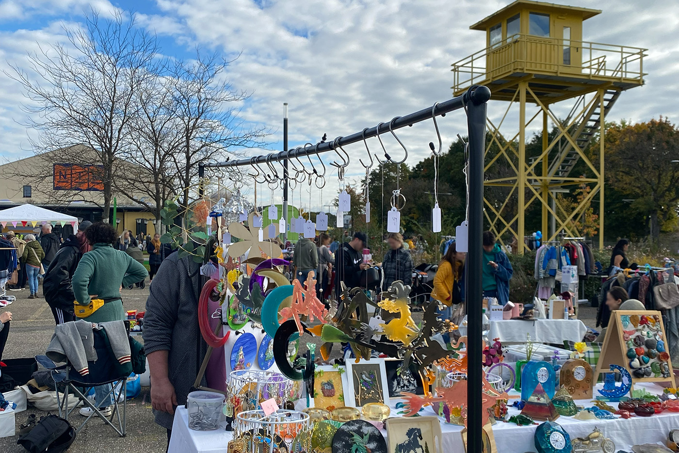 KI-basierte Bildbeschreibung | Markt im Freien mit Tischen, auf denen buntes Kunsthandwerk ausgestellt ist; die Leute stöbern unter einem bewölkten Himmel, mit einem gelben Aussichtsturm im Hintergrund.