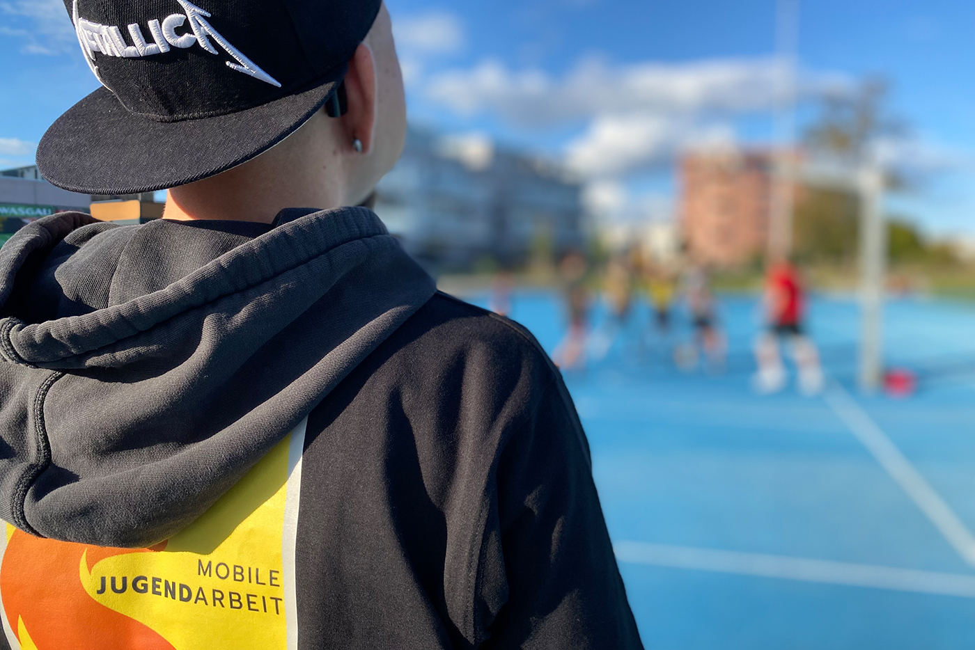 KI-basierte Bildbeschreibung | Eine Person, die eine Metallica-Mütze und eine Jacke mit der Aufschrift Mobile Jugendarbeit trägt, beobachtet eine Gruppe, die auf einem Sportplatz im Freien spielt.