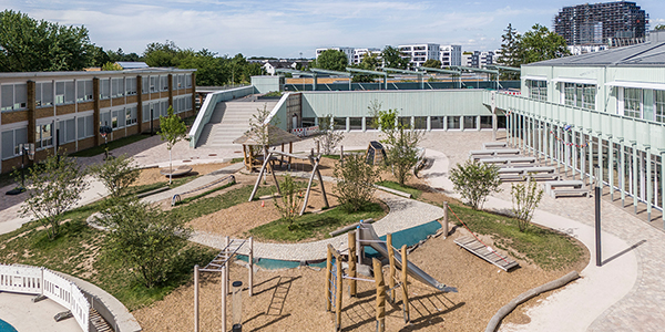 KI-basierte Bildbeschreibung | Ein Schulhof mit einem Spielplatz mit hölzernen Klettergerüsten, umgeben von modernen Gebäuden und Bänken.
