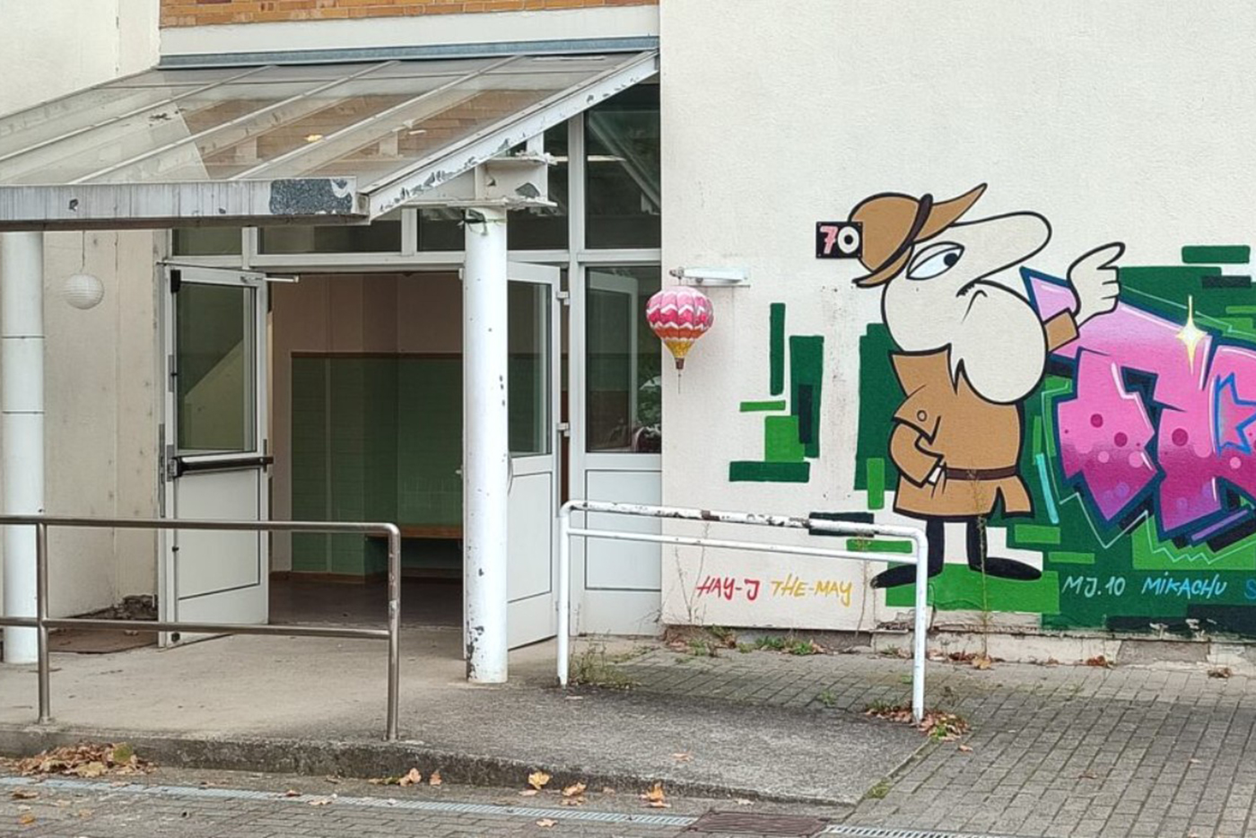 KI-basierte Bildbeschreibung | Auf einer Gebäudewand neben einem offenen Eingang mit Glastüren und einer Rampe ist ein Cartoon-Wandbild eines Detektivs in einem braunen Mantel und Hut gemalt.
