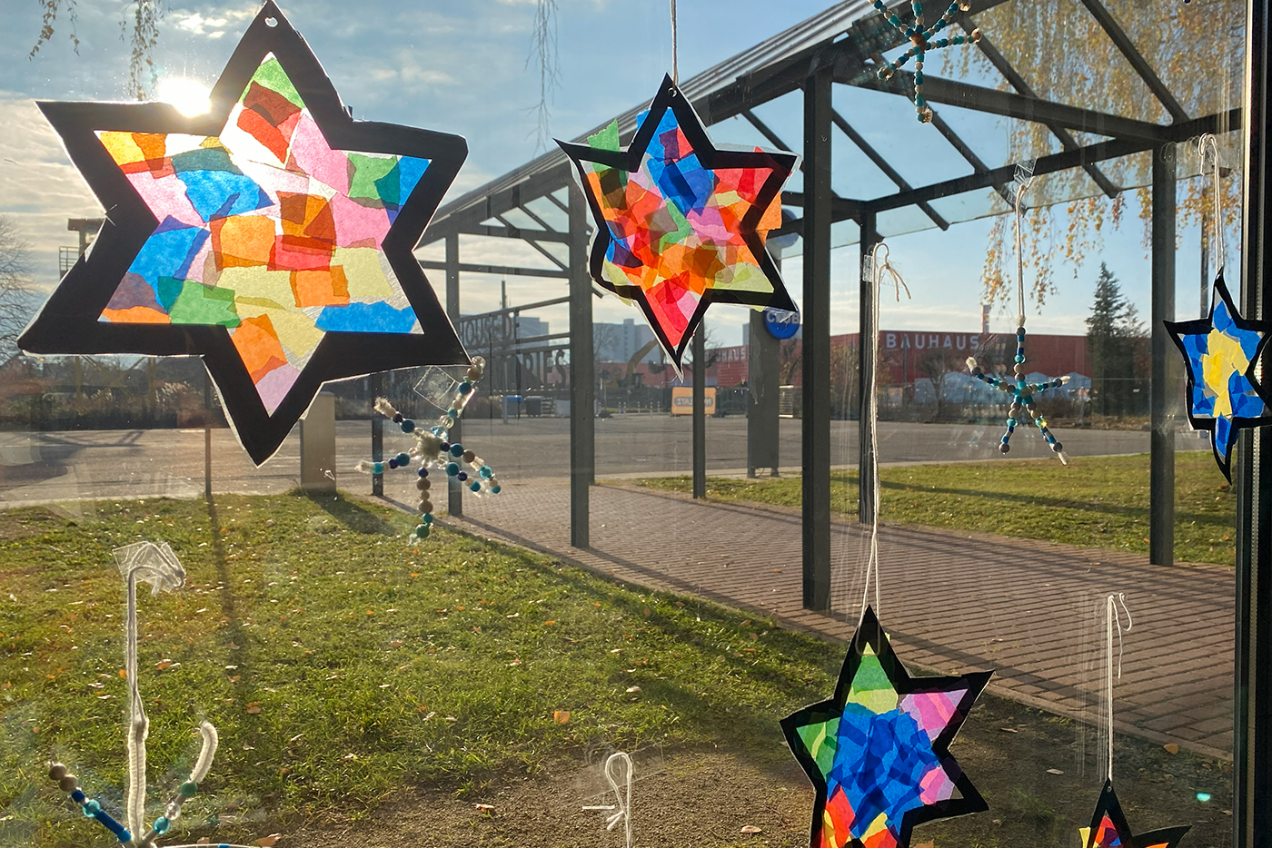 KI-basierte Bildbeschreibung | Bunte sternförmige Sonnenfänger aus Seidenpapier hängen in einem Fenster, durch das Sonnenlicht scheint und im Hintergrund ist ein Bauhaus-Geschäft zu sehen.