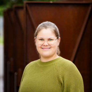 KI-basierte Bildbeschreibung | Eine Person mit Brille und grünem Pullover steht im Freien und lächelt in die Kamera, während im Hintergrund eine Metallkonstruktion zu sehen ist.