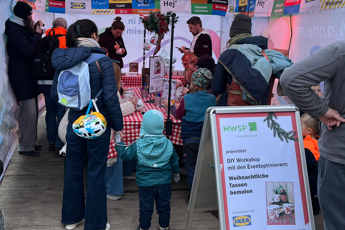KI-basierte Bildbeschreibung | An einem DIY-Workshop-Stand für das Bemalen von Weihnachtstassen versammeln sich Kinder und Erwachsene um Tische mit rot karierten Tüchern. Ein Schild weist auf den Workshop hin.