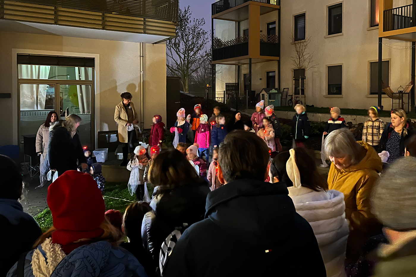 KI-basierte Bildbeschreibung | Eine Gruppe von Kindern und Erwachsenen versammelt sich nachts vor Wohnhäusern, einige Kinder tragen Weihnachtsmannmützen und haben Laternen in der Hand.