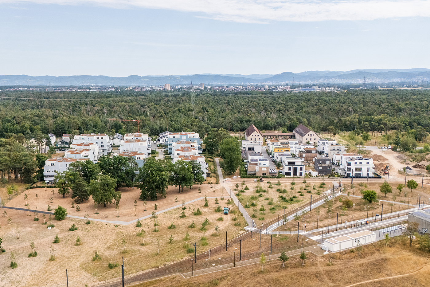 KI-basierte Bildbeschreibung | Luftaufnahme eines Wohngebiets mit modernen Wohngebäuden, umgeben von Bäumen und offenen Flächen, mit einer Stadt und Bergen im Hintergrund.