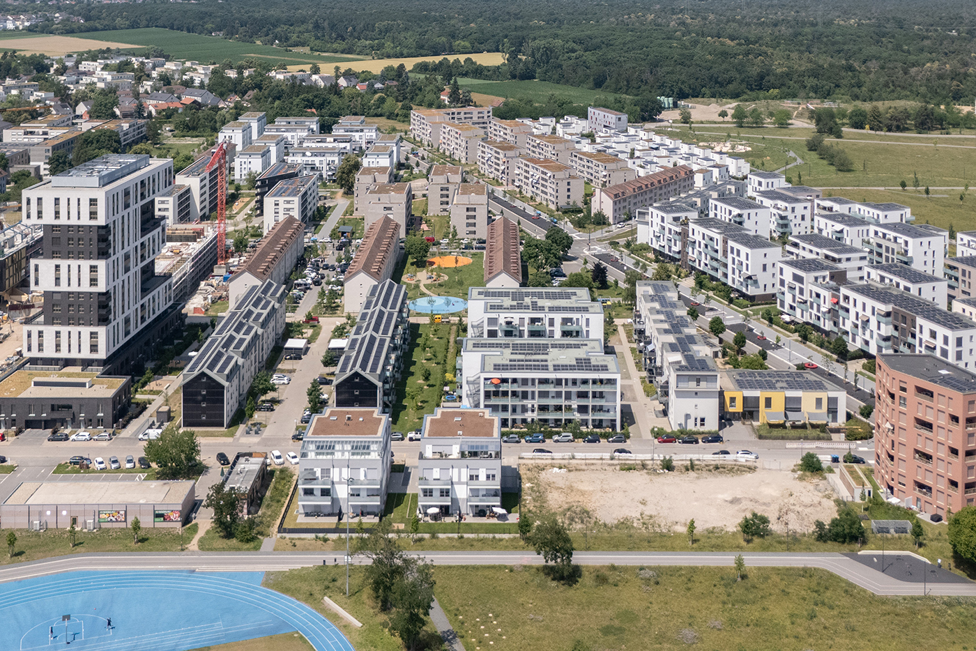 KI-basierte Bildbeschreibung | Luftaufnahme eines modernen Wohn- und Geschäftsviertels mit Wohnhäusern, von Bäumen gesäumten Straßen, Grünflächen und den umliegenden Feldern und Wäldern im Hintergrund.