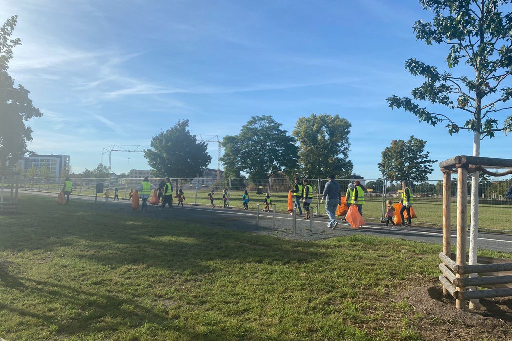 KI-basierte Bildbeschreibung | Eine Gruppe von Kindern und Erwachsenen, die Sicherheitswesten tragen, gehen an einem sonnigen Tag mit orangefarbenen Taschen einen gepflasterten Weg in einem Park entlang, wahrscheinlich im Rahmen eines geplanten Termins oder einer Gemeinschaftsveranstaltung.