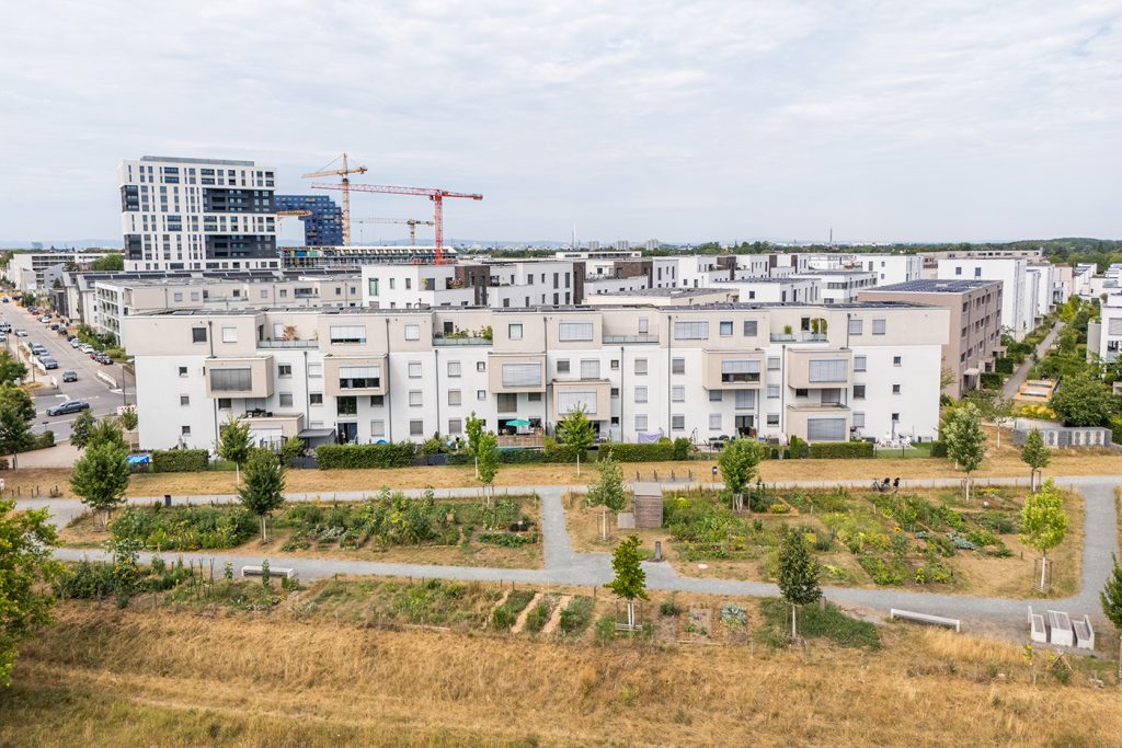 KI-basierte Bildbeschreibung | Eine Reihe moderner Wohnhäuser mit Balkonen liegt gegenüber einem Park mit Spazierwegen, Gärten und Bäumen; im Hintergrund sind Baukräne und weitere Gebäude zu sehen.
