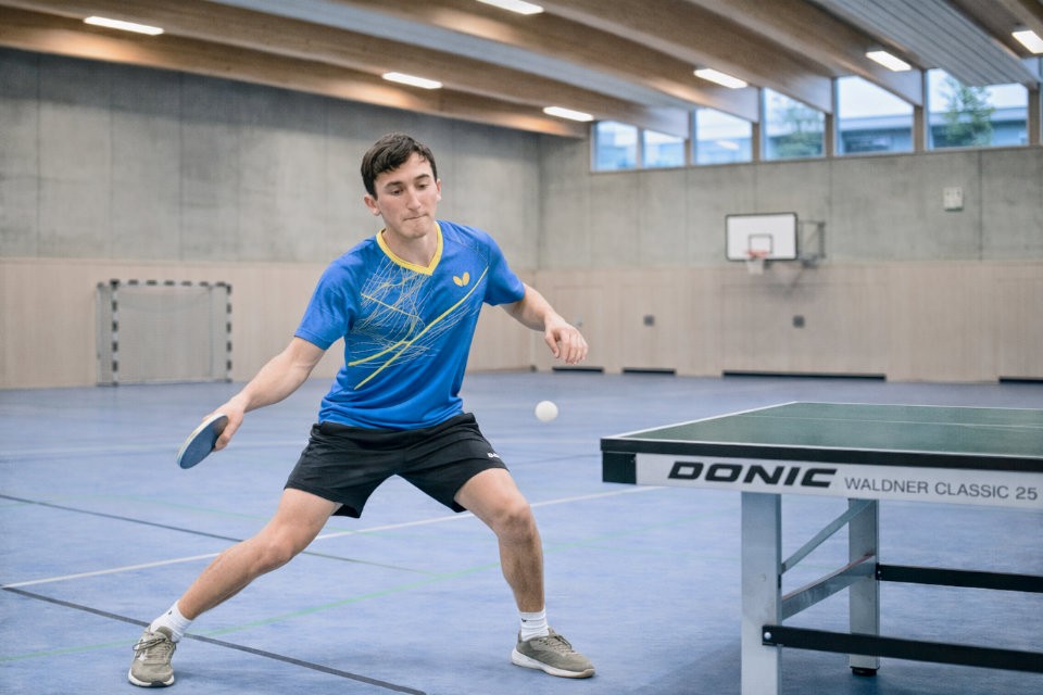 KI-basierte Bildbeschreibung | Ein junger Mann in Sportkleidung spielt Tischtennis in einer Turnhalle und bereitet sich darauf vor, den Ball mit seinem Schläger zu schlagen.