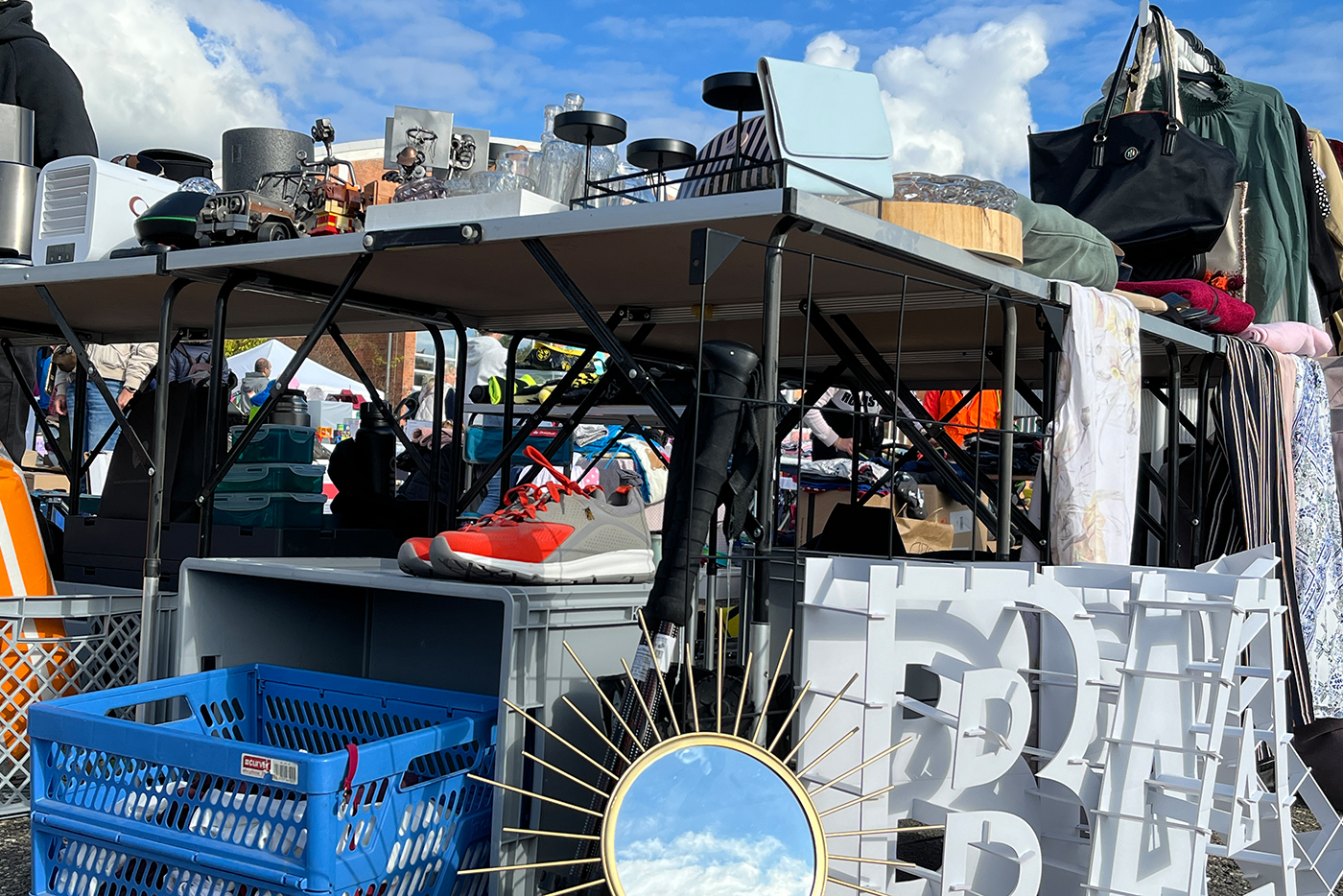 KI-basierte Bildbeschreibung | An einem Flohmarktstand werden verschiedene Gegenstände ausgestellt, darunter ein Paar Turnschuhe, eine blaue Kiste, ein dekorativer Spiegel, elektronische Geräte, Kleidung und Taschen - für jeden Besucher der kommenden Termine unter einem teilweise bewölkten Himmel ist etwas Einzigartiges dabei.