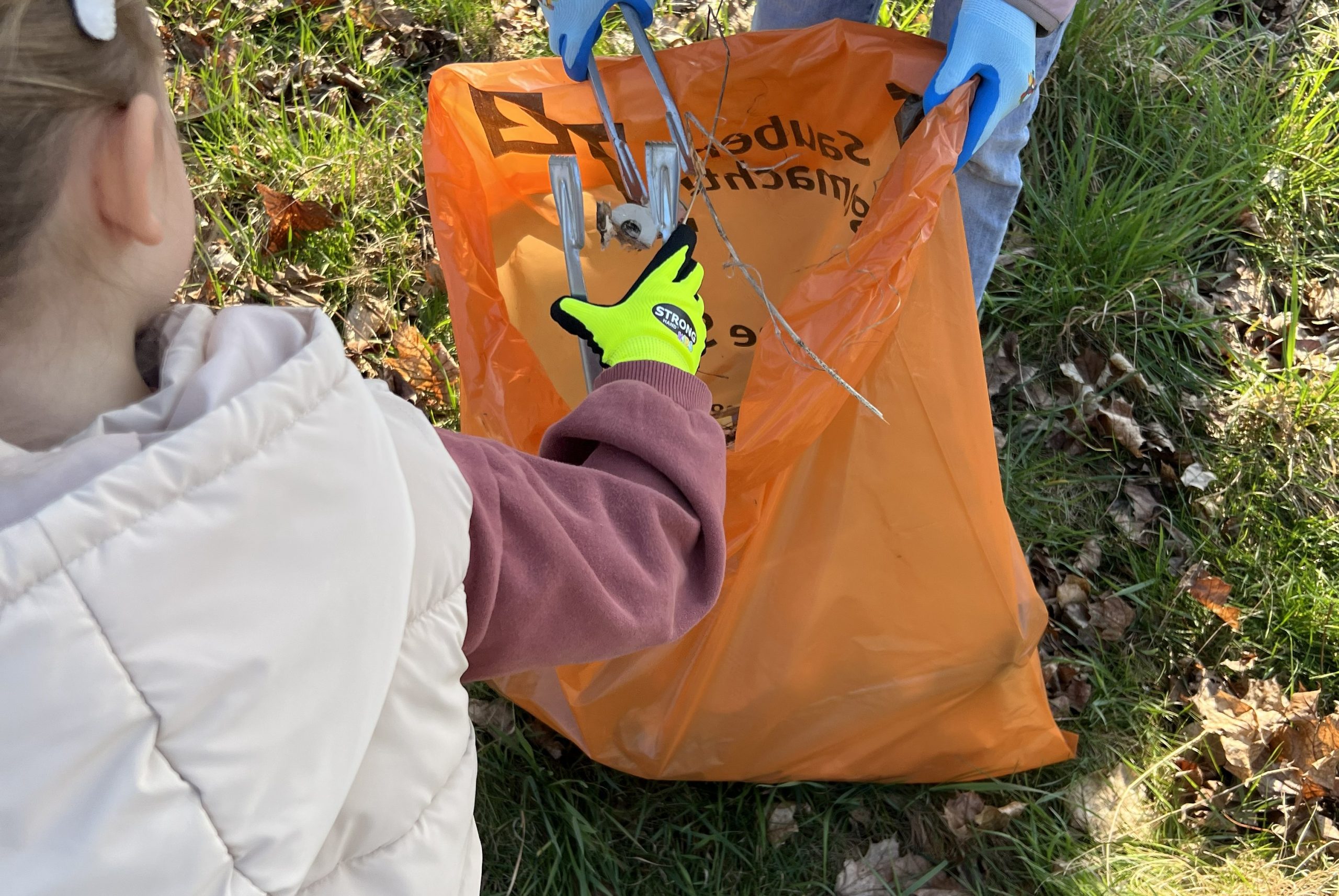 KI-basierte Bildbeschreibung | Ein Kind mit gelben Handschuhen benutzt einen Greifer, um Abfall in einen orangefarbenen Müllsack zu werfen, den eine andere Person auf einer Wiese im Freien hält.
