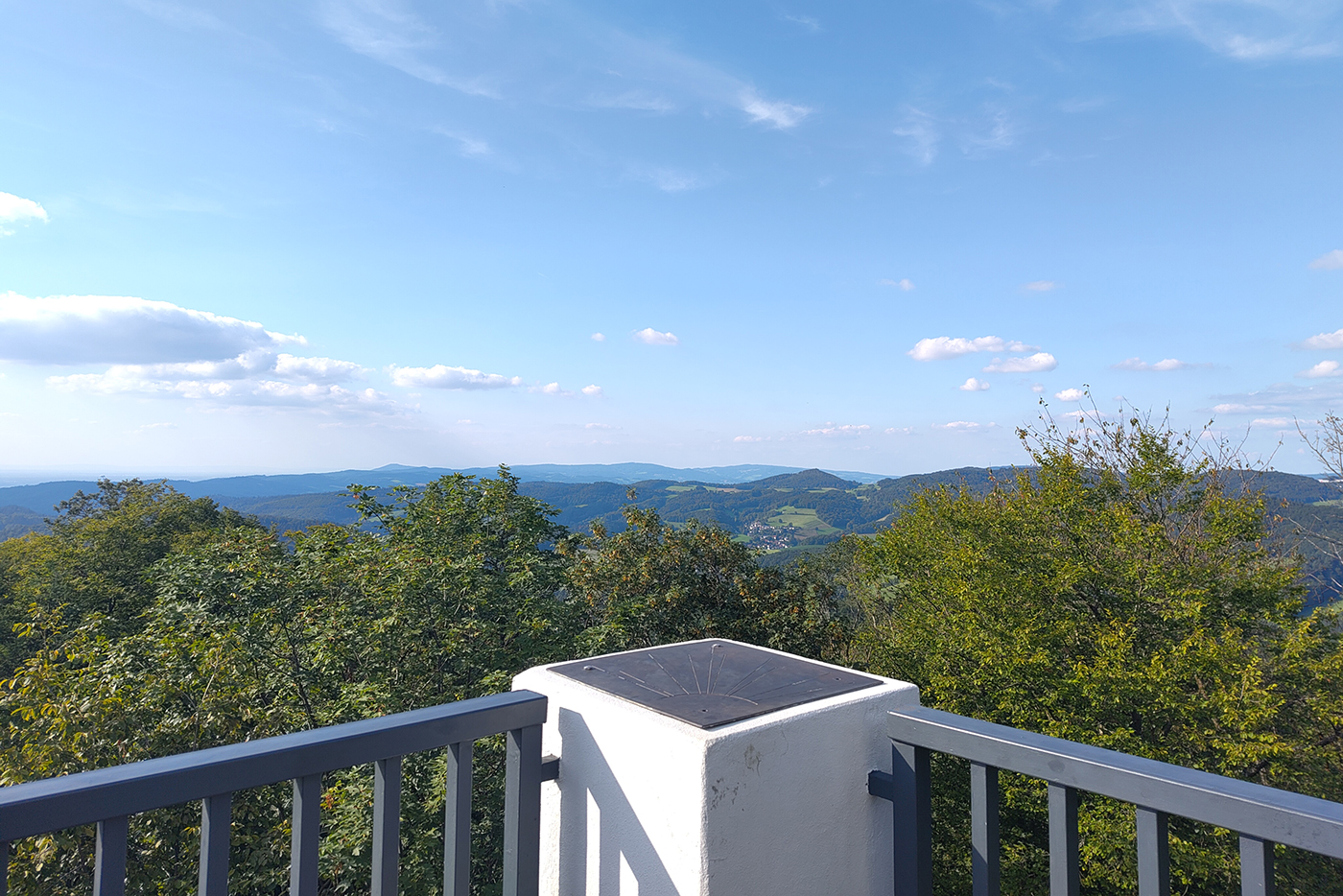 KI-basierte Bildbeschreibung | Ein Aussichtspunkt mit Geländer überblickt die grünen Hügel von Termine, wo vereinzelte Wolken unter einem strahlend blauen Himmel treiben.