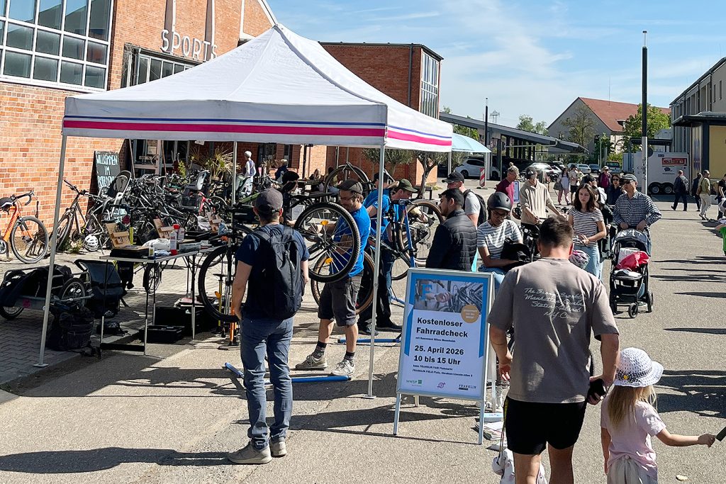 KI-basierte Bildbeschreibung | Menschen versammeln sich um ein weißes Zelt bei einer Fahrradreparaturveranstaltung im Freien. Mehrere Fahrräder werden repariert, während andere in der Schlange warten. An der Stirnseite hängt ein Plakat mit Informationen zur Veranstaltung.