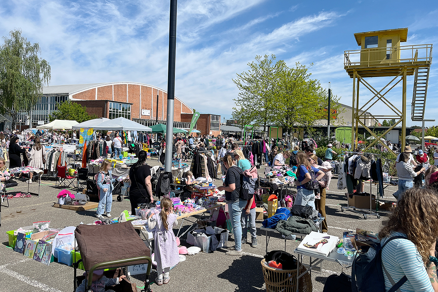 KI-basierte Bildbeschreibung | Flohmarkt im Freien mit Menschen, die an Tischen mit Kleidung, Spielzeug und anderen Gegenständen stöbern; sonniger Tag mit Gebäuden und einem gelben Wachturm im Hintergrund.