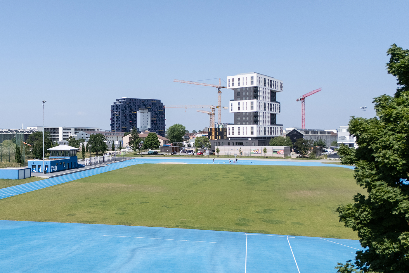KI-basierte Bildbeschreibung | Ein grüner Sportplatz mit einer blauen Laufbahn, mehrere Menschen, die spazieren gehen, moderne Gebäude und Baukräne im Hintergrund unter einem klaren Himmel.