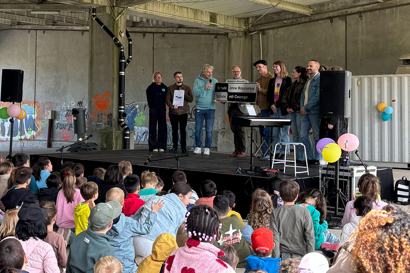 KI-basierte Bildbeschreibung | Eine Gruppe von Menschen steht auf einer Bühne und hält Schilder in die Höhe, während sie sich an ein Publikum von Kindern wendet, die in einem dekorierten Innenraum auf dem Boden sitzen.