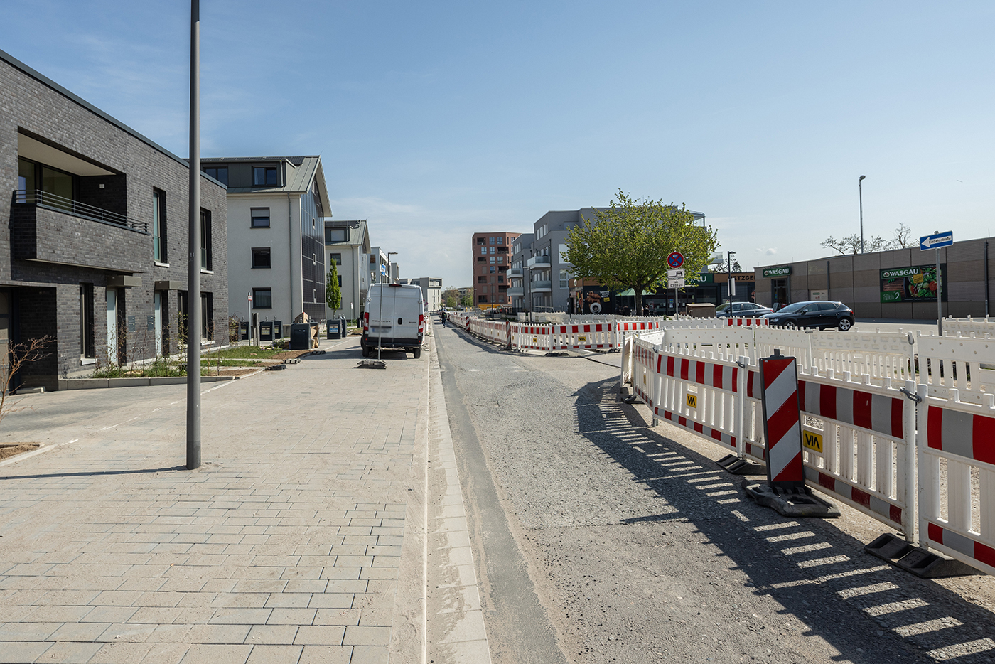 KI-basierte Bildbeschreibung | Eine im Bau befindliche Stadtstraße mit Baustellenabsperrungen, einem geparkten weißen Lieferwagen und modernen Gebäuden auf beiden Seiten bei klarem Himmel.