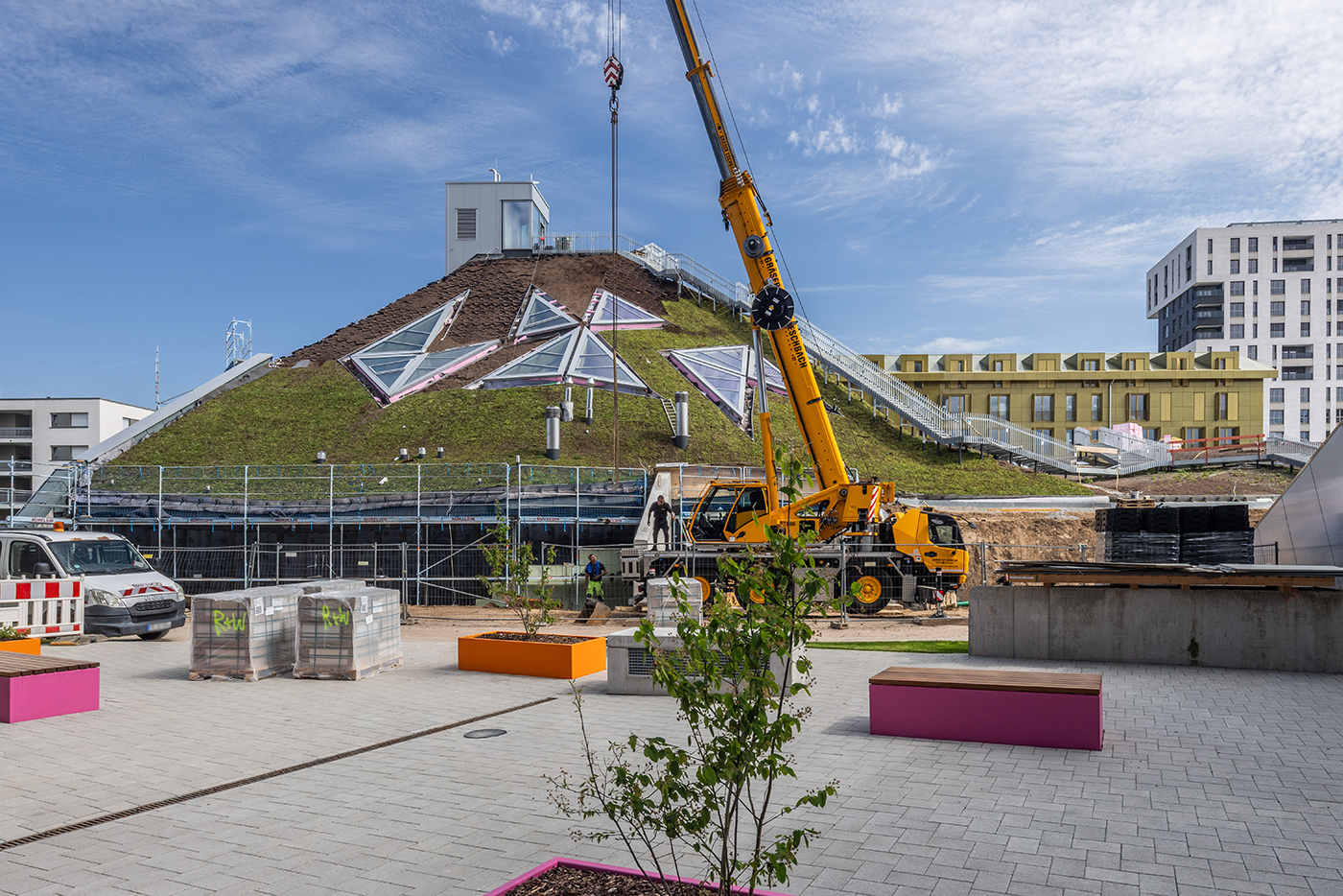 KI-basierte Bildbeschreibung | Ein Kran arbeitet in der Nähe eines grasbewachsenen, pyramidenförmigen Gebäudes, das Investoren anzieht. Umgeben von Baugerüsten, Fahrzeugen und modernen städtischen Strukturen herrscht an einem sonnigen Tag reges Treiben auf der Baustelle.