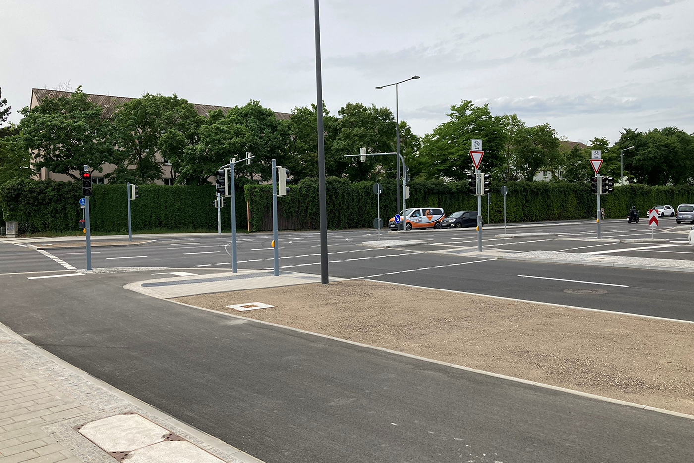 KI-basierte Bildbeschreibung | Eine breite, leere Kreuzung mit Ampeln, Vorfahrtsschildern und ein paar Autos und Fußgängern im Hintergrund unter einem bewölkten Himmel.