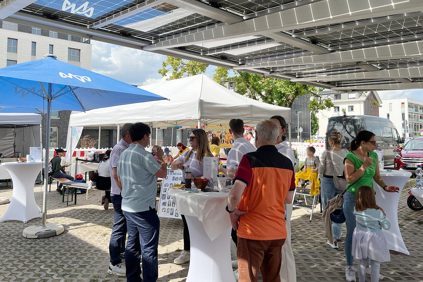 KI-basierte Bildbeschreibung | Menschen versammeln sich und unterhalten sich unter Zelten und einem Solarmodul-Dach bei einer Veranstaltung im Freien, mit Informationstischen, Sonnenschirmen und Kindern.