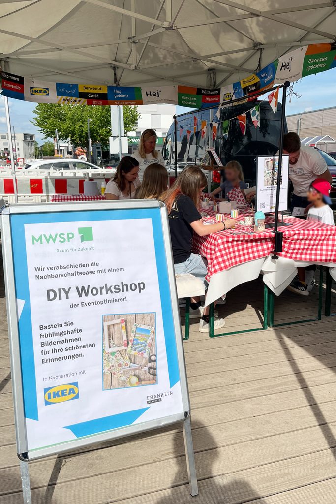 KI-basierte Bildbeschreibung | Menschen nehmen an einem DIY-Bilderrahmen-Workshop bei einer Freiluftveranstaltung unter einem Zelt teil, mit einem Schild, das für die Aktivität in Partnerschaft mit IKEA und Franklin wirbt.