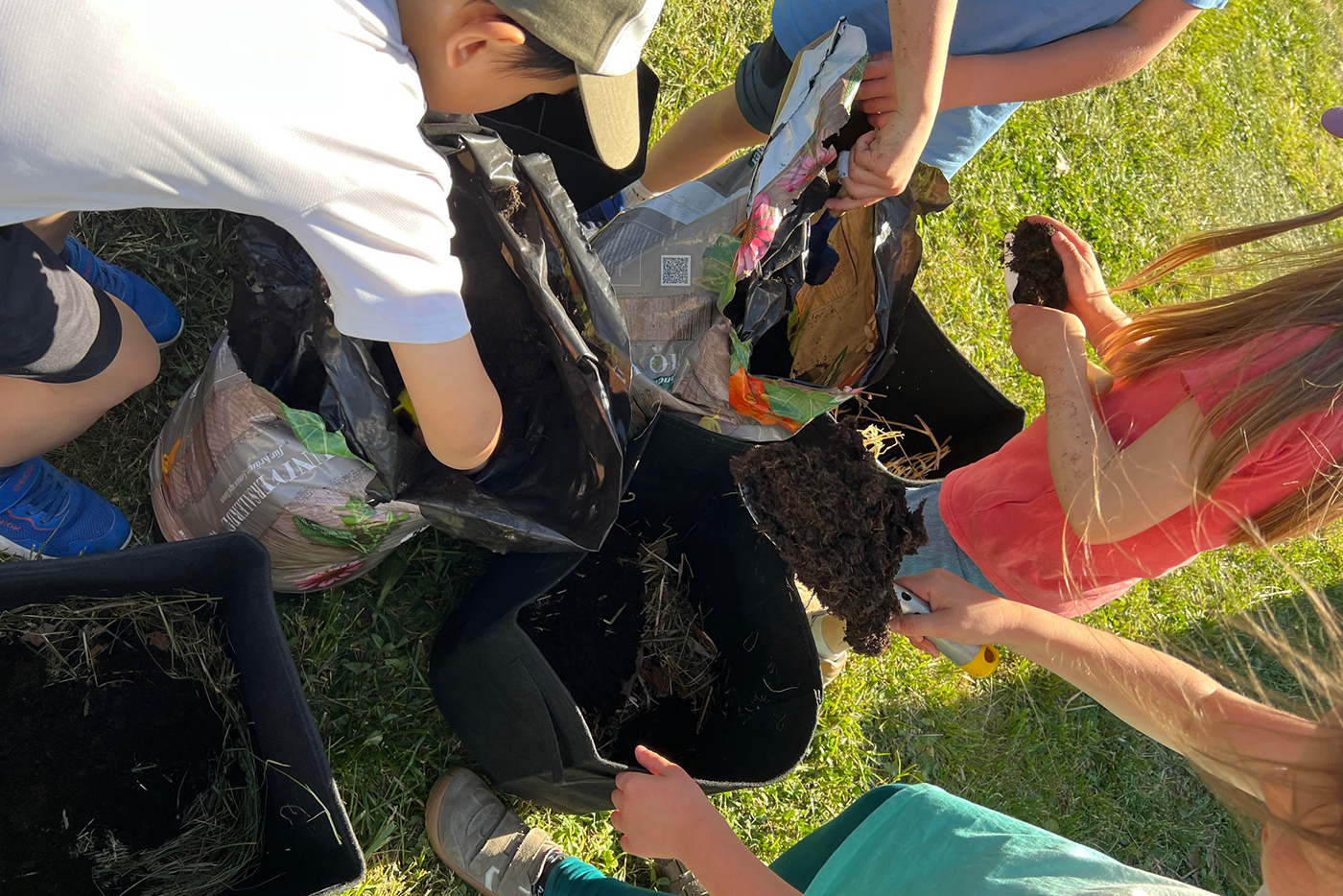 KI-basierte Bildbeschreibung | Vier Kinder füllen auf einer Wiese schwarze Stofftöpfe mit Erde und Kompost.