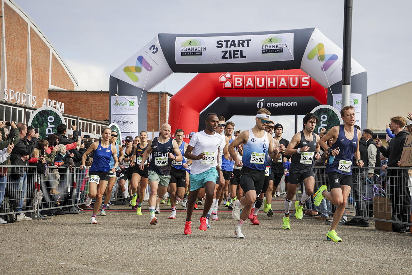 KI-basierte Bildbeschreibung | Läufer in Sportbekleidung starten bei einer Veranstaltung im Freien in ein Rennen und laufen unter einem aufblasbaren Bogen mit der Aufschrift „START ZIEL“ hindurch, während Zuschauer und Organisatoren hinter Absperrungen ihre Termine überprüfen.