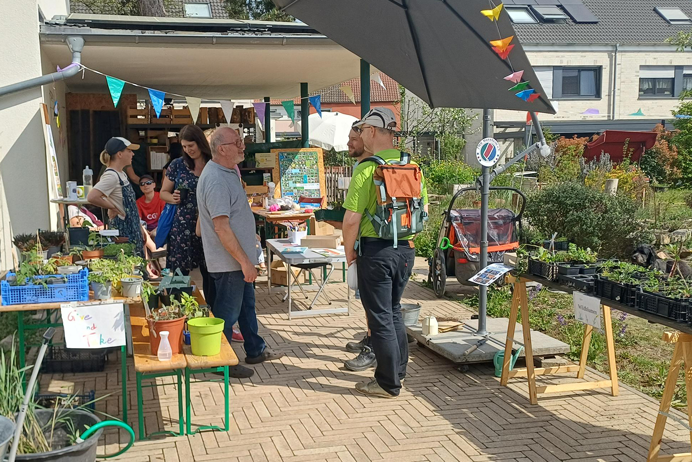 KI-basierte Bildbeschreibung | Menschen versammeln sich bei einem Pflanzenverkauf im Freien, stöbern und unterhalten sich an Tischen mit Topfpflanzen unter Sonnenschirmen und festlichen Wimpeln, während im Hintergrund Häuser zu sehen sind.