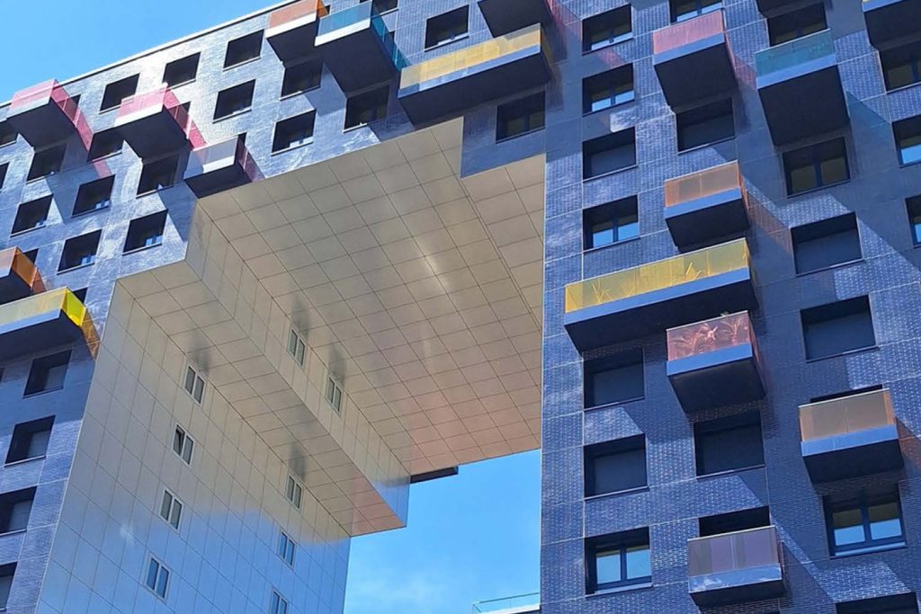 KI-basierte Bildbeschreibung | Ein modernes Gebäude mit einem zentralen, erhöhten Durchgang, kleinen, farbenfrohen Balkonen und einer blau gekachelten Fassade vor dem Hintergrund eines klaren Himmels – perfekt für auffällige „Termine“-Räume.