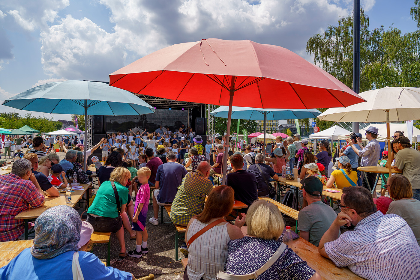 KI-basierte Bildbeschreibung | An einem sonnigen Tag bei einem Festival oder einer Veranstaltung sitzt und steht eine Menschenmenge unter großen Sonnenschirmen und verfolgt eine Aufführung auf einer Freiluftbühne.