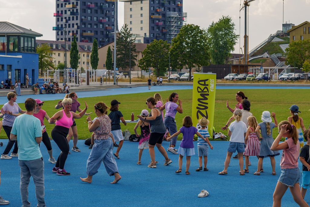KI-basierte Bildbeschreibung | Erwachsene und Kinder nehmen an einem Zumba-Gruppenkurs im Freien auf einer blauen Laufbahn teil, im Hintergrund sind Gebäude und geparkte Autos zu sehen.