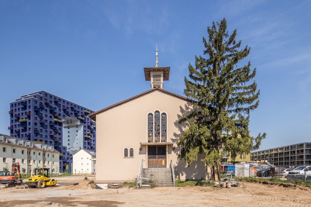 KI-basierte Bildbeschreibung | Hinter einer großen Kiefer steht eine beigefarbene Kirche mit Buntglasfenstern, umgeben von Baumaschinen und modernen Wohngebäuden unter einem strahlend blauen Himmel.