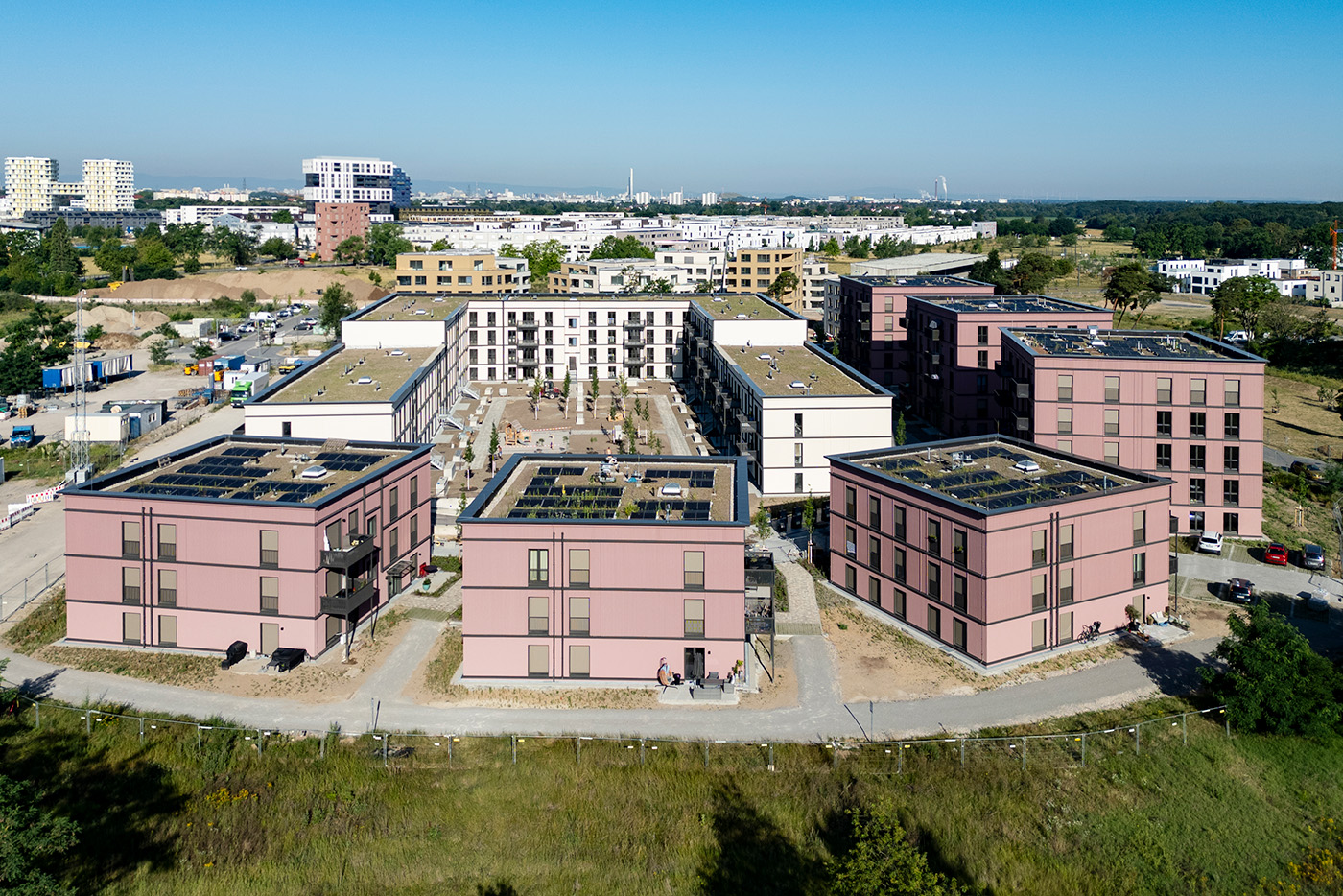 KI-basierte Bildbeschreibung | Luftaufnahme eines modernen Wohnkomplexes – oder „Quartiere“ – mit mehreren rechteckigen, rosafarbenen Gebäuden, begrünten Dächern und umliegenden Wegen inmitten eines städtischen Gebiets.