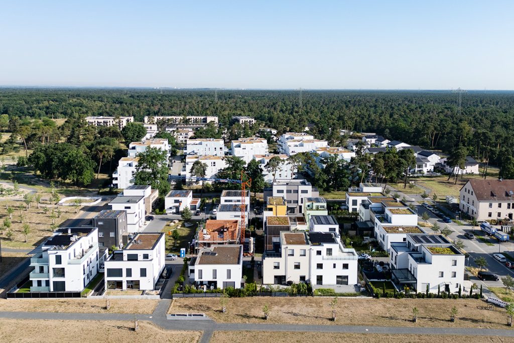 KI-basierte Bildbeschreibung | Luftaufnahme eines modernen Wohnviertels mit weißen mehrstöckigen Häusern, grünen Bäumen und einem Wald im Hintergrund unter klarem Himmel.
