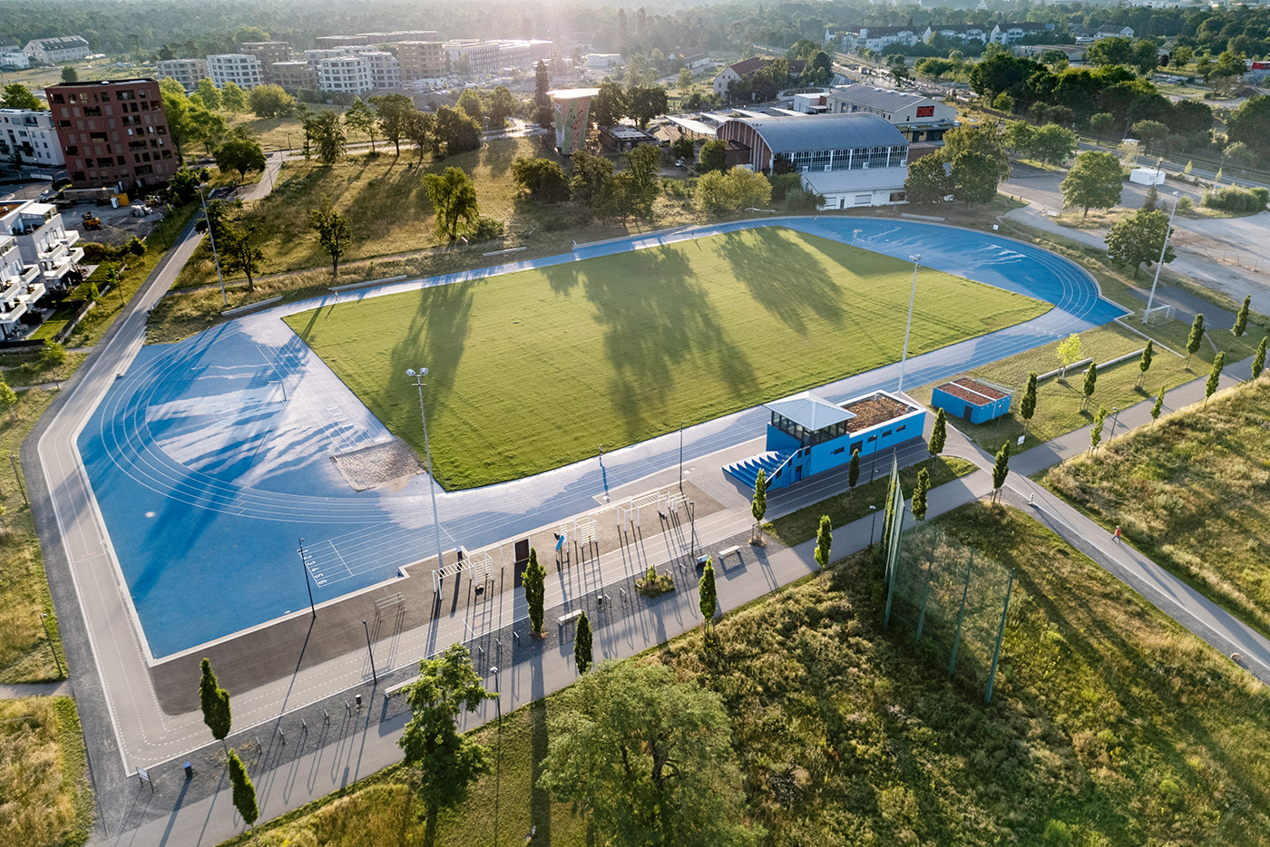 KI-basierte Bildbeschreibung | Luftaufnahme eines Freiluft-Sportstadions mit einem leuchtend grünen Rasenfeld, einer blauen Laufbahn, einer Zuschauertribüne sowie umliegenden Bäumen und Gebäuden – eine dynamische Kulisse, die den Geist des Sports verkörpert.
