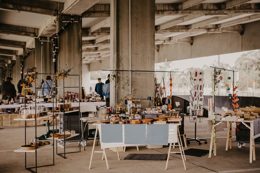KI-basierte Bildbeschreibung | An den Ständen des überdachten Marktes werden verschiedene handgefertigte Waren, Töpferwaren und Kunsthandwerk angeboten. Im Hintergrund sind unter einem großen Betonbau einige Menschen zu sehen, die nach einzigartigen Fundstücken und den nächsten Terminen Ausschau halten.