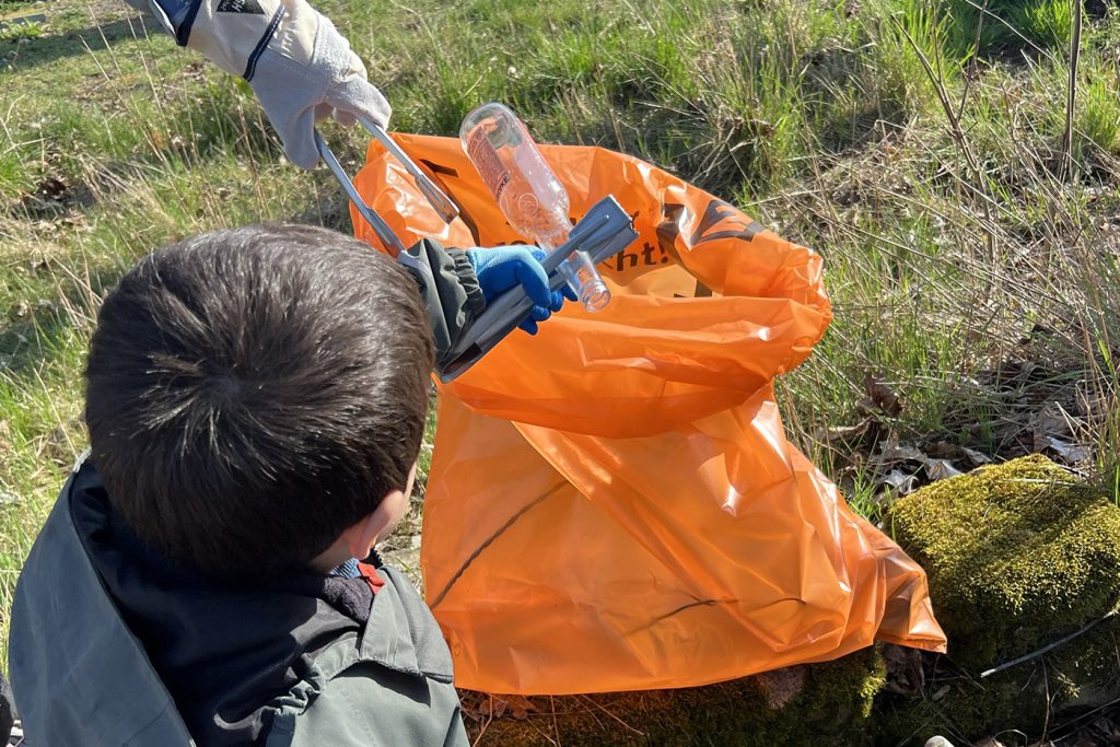 KI-basierte Bildbeschreibung | Kinder und Erwachsene verwenden Greifzangen, um Plastikflaschen aufzuheben und sie draußen auf einer Rasenfläche in einen orangefarbenen Müllsack zu legen, und tragen so dazu bei, die Umwelt während der „Termine“ sauber zu halten.